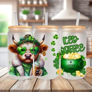 Op de afbeelding: Drie frosted glazen bekers met deksels en rietjes. De beker links heeft een cartoon-koe die een groen-witte hoed en zonnebril draagt. De beker in het midden is leeg. De beker rechts heeft de tekst "Iced Coffee & Lucky Vibes" met een pot goud en klavertjesvier.