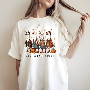 Op de afbeelding: Wit T-shirt met een illustratie van vier ganzen in herfstoutfits, inclusief hoeden en vesten, met pompoenen. De tekst "JUST A FALL GOOSE" staat eronder. Het ontwerp heeft een seizoensgebonden thema.