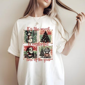 Op de afbeelding: Wit T-shirt met een kerstthema. Het ontwerp toont een sneeuwpop, kerstboom, kerstman en een Schotse Hooglander, met de tekst "It's the most Wonderful time of the year."