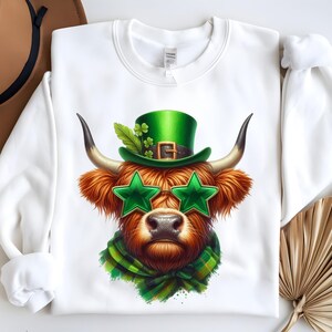 Op de afbeelding: Wit sweatshirt met een afbeelding van een Schotse hooglander die een groene leprechaun-hoed en zonnebril draagt. De koe draagt een groen-witte geruite sjaal.