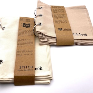 May include: Two fabric sketchbooks with brown paper covers. The covers have the text "STITCH Fabric Sketchbook" printed on them. The sketchbooks are bound with silver rings. The sketchbooks are on a white surface.