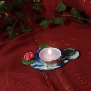 Può includere: Una candela tealight accesa in un supporto argentato è posizionata su una base decorativa in ceramica. La base è bianca con motivi blu e bianchi e presenta un fiore rosa e ninfee verdi. Lo sfondo è un tessuto rosso.