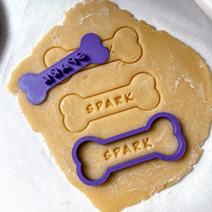 Peut inclure: Emporte-pièces en forme d'os de chien en plastique violet avec les mots "WAG" et "SPARK" dessus. Les emporte-pièces sont placés sur une feuille de pâte à biscuits non cuite.