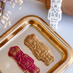 May include: A gold-coloured metal tray holds two decorated biscuits shaped like nutcrackers, one in burgundy and gold, the other in gold. A white plastic nutcracker biscuit cutter and a wooden rolling pin are also in the image.
