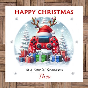 Puede incluir: Una tarjeta de Navidad con el texto "HAPPY CHRISTMAS" en rojo. Un mando de juego rojo con un gorro de Papá Noel y cuernos de reno está en el centro. La tarjeta dice "To a Special Grandson Theo" y presenta regalos envueltos.