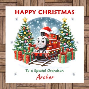 Puede incluir: Una tarjeta de felicitación navideña cuadrada con una locomotora de vapor roja de dibujos animados con gorro de Papá Noel, entre dos árboles de Navidad decorados y regalos. El texto dice HAPPY CHRISTMAS, To a Special Grandson y Archer.