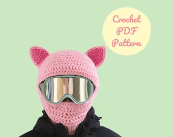 Op de afbeelding: Roze gebreide skibril met oorflappen en een gespiegelde skibril. De tekst "Crochet PDF Pattern" staat in een cirkel op een lichtgroene achtergrond.