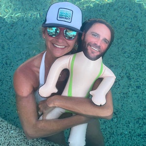 Pode incluir: Uma mulher em uma piscina usando um chapéu branco com o texto "slande77" nele, segurando um flutuador de piscina inflável branco em forma de pessoa vestindo um colete verde.