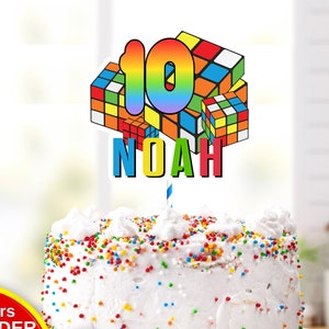 Op de afbeelding: Een wit glazuurcake met kleurrijke hagelslag, bekroond met een caketopper met een regenboogkleurig nummer 10 en de naam "NOAH" in blauw. De cake staat op een wit bord.