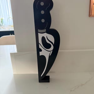 Puede incluir: Cuchillo decorativo de madera negra con un diseño gráfico blanco. El cuchillo tiene un mango con tres detalles circulares y una hoja con una cara estilizada. El cuchillo está de pie sobre una pequeña base negra.