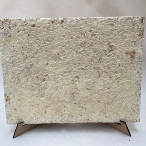 Puede incluir: Una pieza de arte de papel rectangular, texturizada y blanca, sobre un soporte de madera. El papel tiene un aspecto rugoso y hecho a mano.