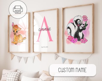 May include: Three framed art prints with a nursery theme. One features a fawn, another a pink letter "A" with the name "annie" and date, and the third a skunk holding a flower. The prints have watercolor backgrounds and are in light wood frames.
