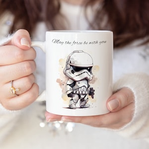 Può includere: Tazza in ceramica bianca con un'illustrazione di un soldato imperiale di Star Wars in stile cartone animato. La tazza ha la scritta "Che la forza sia con te" stampata su di essa.
