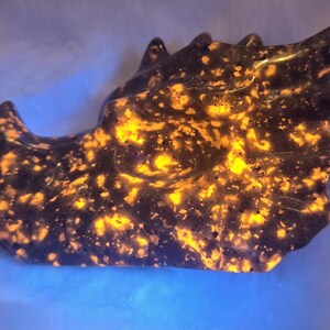May include: A dark, sculpted stone in the shape of a dragon's head. The stone is primarily black with vibrant orange and yellow inclusions, creating a speckled, glowing effect. The dragon's head is detailed with horns and a defined snout.