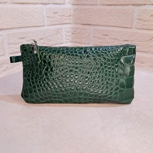 May include: A rectangular dark green pouch featuring a glossy, embossed crocodile skin texture. The accessory includes a silver-toned metal zipper closure along the top edge, a small side tab, and a thin green leather pull attached to the zipper slider.