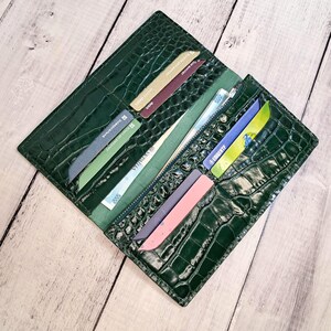 Peut inclure: Un portefeuille vert fonc&eacute; avec un motif textur&eacute; en peau de crocodile. L'int&eacute;rieur pr&eacute;sente plusieurs fentes pour cartes contenant diverses cartes color&eacute;es, un compartiment central zipp&eacute; et une longue poche pour les billets de banque.