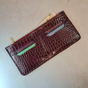 May include: A burgundy leather wallet with a crocodile skin texture. The wallet has a gold zip and a red interior. Several cards and a banknote are visible inside the wallet.