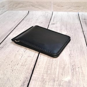May include: A black leather money clip wallet with a silver clip. The rectangular wallet has stitched edges. The background is a light wood grain.