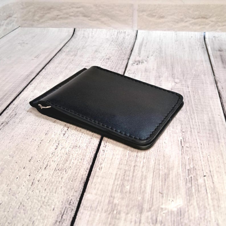 May include: A black leather money clip wallet with a silver clip. The rectangular wallet has stitched edges. The background is a light wood grain.