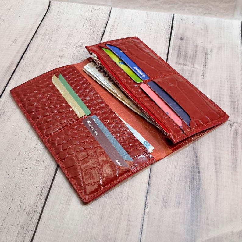 Peut inclure: Un portefeuille rouge avec un motif textur&eacute; en peau de crocodile, dot&eacute; de plusieurs fentes pour cartes et d'un compartiment zipp&eacute;. Le portefeuille est ouvert, r&eacute;v&eacute;lant plusieurs cartes en plastique color&eacute;es et des billets rang&eacute;s dans les sections.
