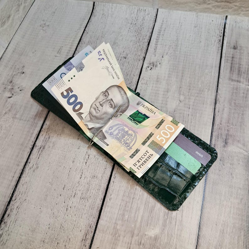 May include: A dark green leather money clip wallet with a crocodile pattern, holding several Ukrainian 500 Hryvnia banknotes and a debit card. The wallet is open, displaying the money and card.