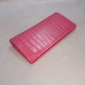 Peut inclure: Un portefeuille rectangulaire d'un rose vif avec un motif textur&eacute; en peau de crocodile. Le portefeuille pr&eacute;sente un design pli&eacute; avec des coutures visibles le long des bords et une finition lisse et uniforme sur toute la surface ext&eacute;rieure.
