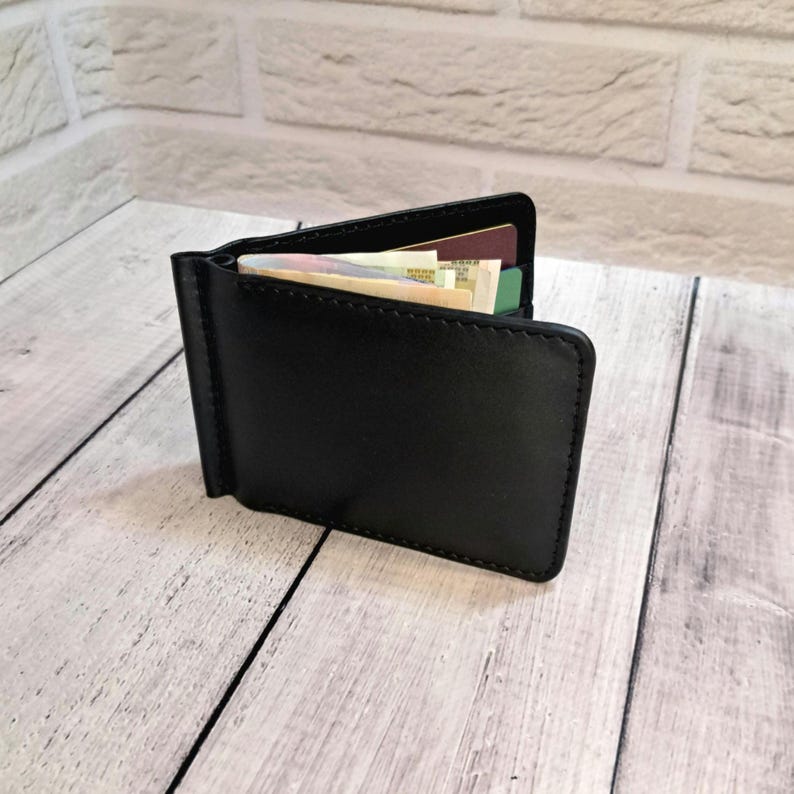 May include: A black leather money clip wallet, open to show banknotes and cards. The wallet has a rectangular shape with rounded corners and a metal clip. The background is a light-coloured wooden surface and a white brick wall.