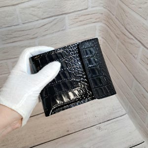 Könnte beinhalten: Eine schwarze, glänzende Geldbörse mit Krokodilhautstruktur. Die Geldbörse ist rechteckig mit einem Klappenverschluss. Das Bild zeigt die Geldbörse, die von einer behandschuhten Hand vor einem hellen Holz- und Ziegelhintergrund gehalten wird.