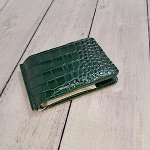 May include: A dark green leather money clip wallet with a crocodile skin texture. The wallet is open, revealing a few banknotes inside. The clip is silver-coloured.