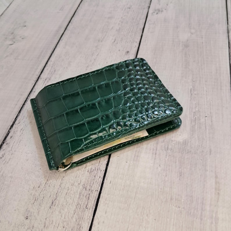 May include: A dark green leather money clip wallet with a crocodile skin texture. The wallet is open, revealing a few banknotes inside. The clip is silver-coloured.