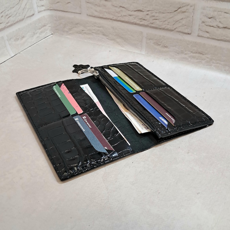 Peut inclure: Un portefeuille noir pliable avec un motif textur&eacute; en peau de crocodile. L'int&eacute;rieur pr&eacute;sente plusieurs fentes pour cartes contenant des cartes de couleurs vari&eacute;es et un compartiment zipp&eacute; avec une tirette en m&eacute;tal argent&eacute;.