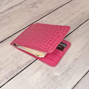 May include: A bright pink bifold wallet featuring a textured crocodile skin pattern. The wallet is open, revealing a metal money clip holding paper currency and a card slot containing a dark card with the visible text debit printed in white.