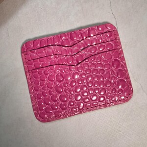 Puede incluir: Un tarjetero de color rosa brillante con un patrón texturizado de piel de cocodrilo. El accesorio rectangular tiene esquinas redondeadas y dos ranuras horizontales para tarjetas, con costuras visibles a lo largo de los bordes del material.
