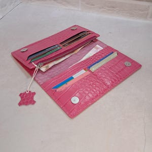 May include: Open pink leather wallet with a crocodile texture. The wallet is open, revealing multiple card slots, a zippered compartment, and cash. A small leather tag hangs from the zipper. The wallet is secured with magnetic snaps.