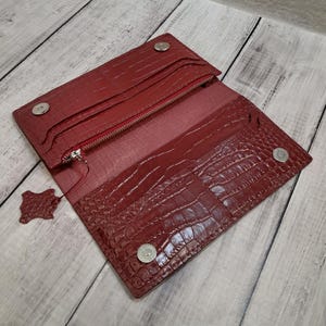 May include: Open, red leather wallet with a crocodile skin texture. The wallet has multiple compartments, a zippered pocket, and snap closures. A small leather swatch is attached. The wallet is open, revealing the interior.