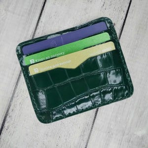 以下が含まれることがあります： クロコダイル柄のダークグリーンのカードケース。紫、緑、金色の3枚のカードが入っています。長方形のカードケースは角が丸みを帯びています。