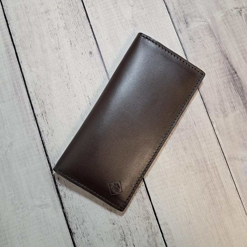 Peut inclure: Un portefeuille long en cuir marron fonc&eacute; avec une finition lisse et mate et des coutures visibles le long des bords. Le portefeuille pr&eacute;sente une forme rectangulaire et un petit logo en forme de losange gaufr&eacute; situ&eacute; pr&egrave;s du coin inf&eacute;rieur.