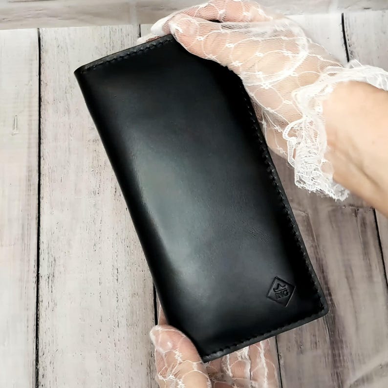 Peut inclure: Un long portefeuille rectangulaire en cuir noir avec des coutures visibles sur les bords. Le portefeuille pr&eacute;sente un petit logo en forme de losange gaufr&eacute; dans le coin inf&eacute;rieur droit, contenant une ic&ocirc;ne de couronne et les lettres ING.