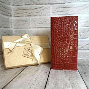 Peut inclure: Un portefeuille rectangulaire rouge fonc&eacute; avec une texture gaufr&eacute;e en peau de crocodile brillante est pos&eacute; debout &agrave; c&ocirc;t&eacute; d'une bo&icirc;te cadeau en carton beige entour&eacute;e d'un ruban cr&egrave;me. Une &eacute;tiquette carr&eacute;e porte le texte ING INGVARR.