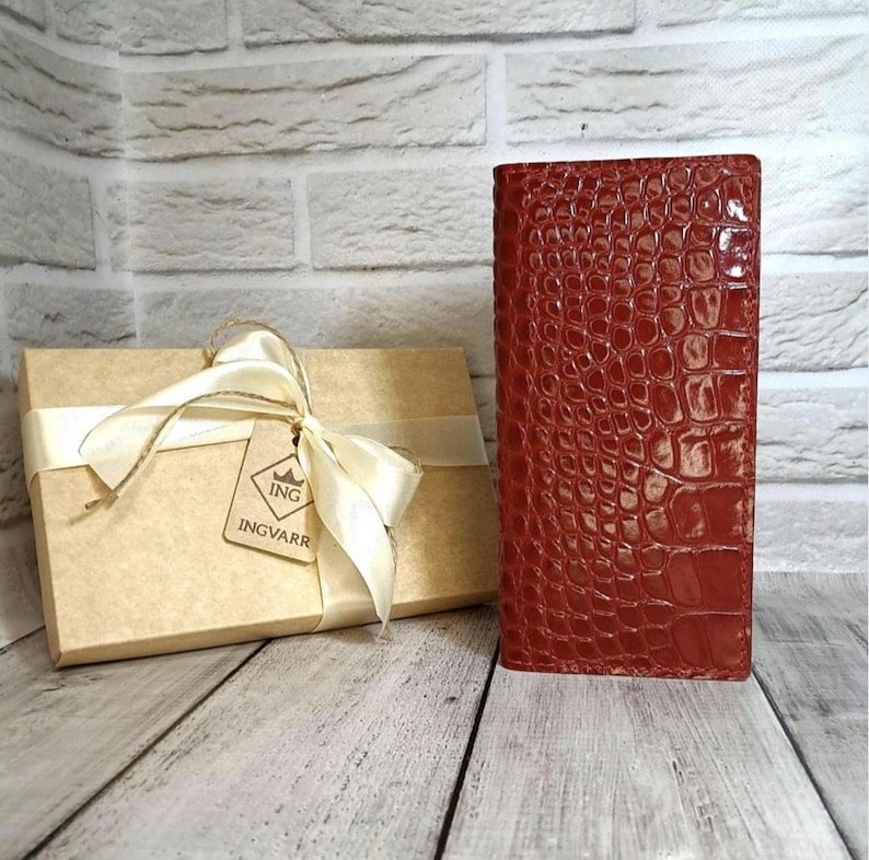 Peut inclure: Un portefeuille rectangulaire rouge fonc&eacute; avec une texture gaufr&eacute;e en peau de crocodile brillante est pos&eacute; debout &agrave; c&ocirc;t&eacute; d'une bo&icirc;te cadeau en carton beige entour&eacute;e d'un ruban cr&egrave;me. Une &eacute;tiquette carr&eacute;e porte le texte ING INGVARR.
