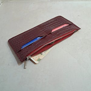 Pode incluir: Uma carteira de couro bordô com um padrão texturizado semelhante ao de um crocodilo. A carteira está aberta, revelando um interior vermelho, um zíper e alguns cartões e dinheiro. O zíper é prateado.