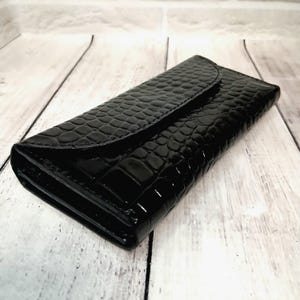 May include: A rectangular black wallet featuring a glossy finish and a textured crocodile skin pattern. The wallet has a flap closure that extends across the top, with visible stitching along the edges and a slim, elongated profile.