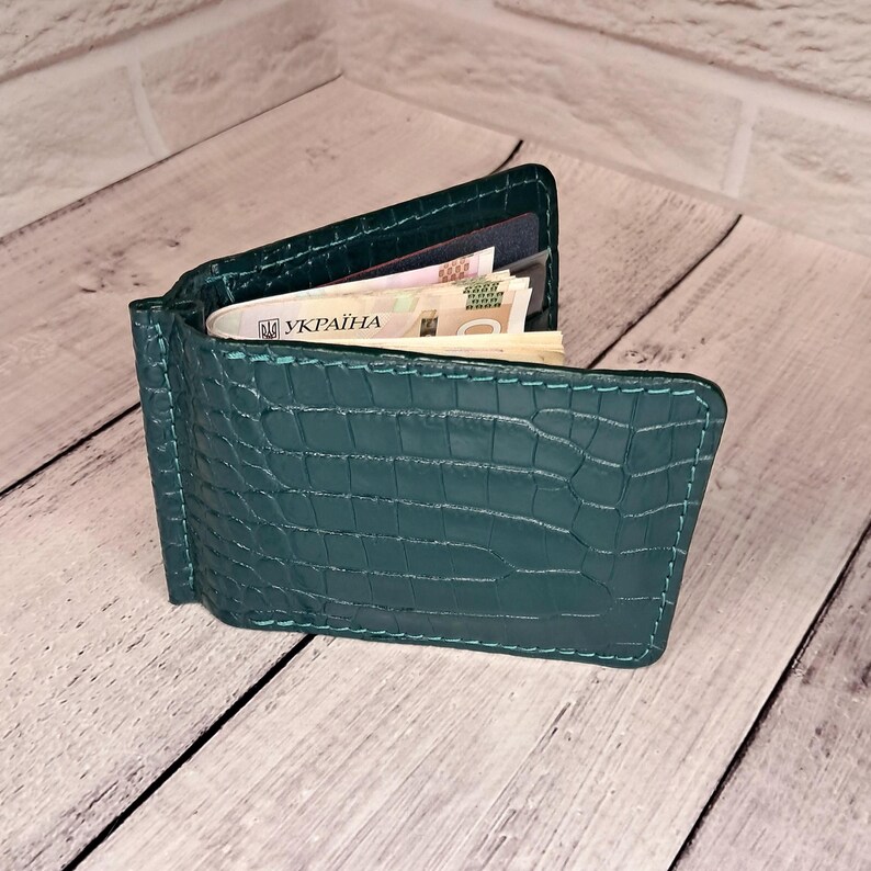 May include: Teal green leather wallet with a crocodile skin texture. The wallet is open, revealing cash and cards. The stitching is a lighter teal colour. The wallet is on a light wood surface.