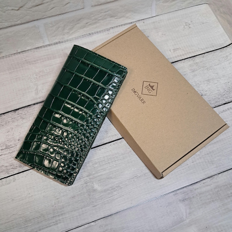 Peut inclure: Un portefeuille rectangulaire vert fonc&eacute; avec une texture brillante fa&ccedil;on peau de crocodile. &Agrave; c&ocirc;t&eacute; du portefeuille se trouve une bo&icirc;te cadeau rectangulaire en carton portant le nom de marque INGVARR imprim&eacute; en noir avec un logo en losange.