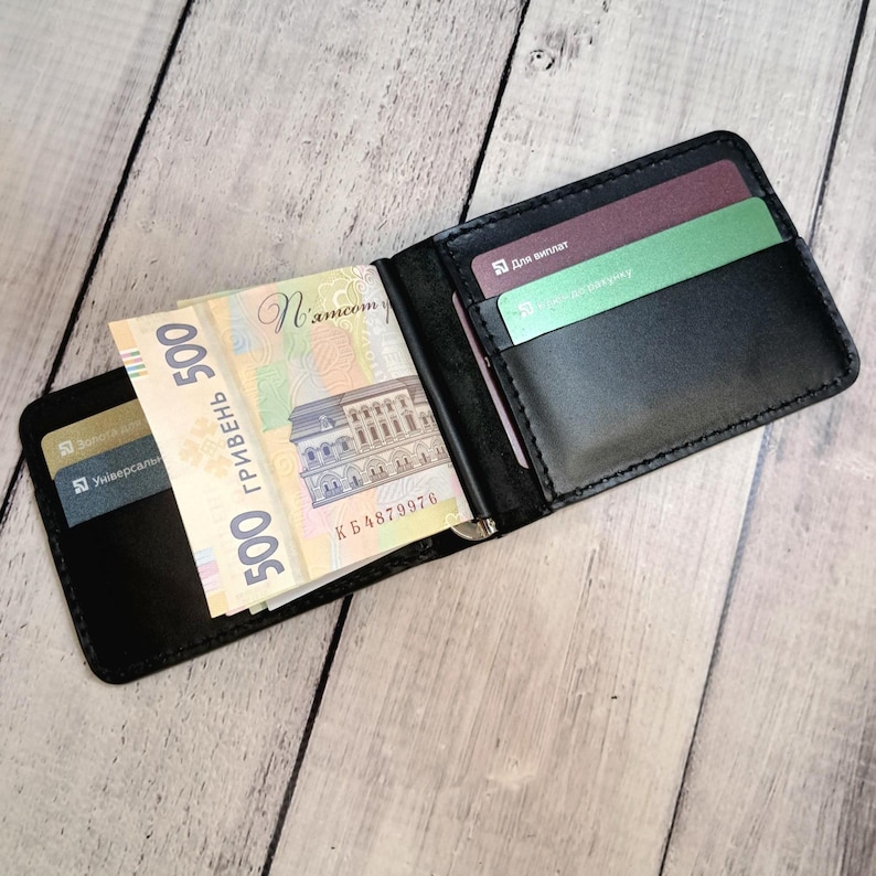 May include: Black leather money clip wallet containing several 500 Ukrainian Hryvnia banknotes and credit cards. The wallet is open, revealing the banknotes held by a metal clip. The background is light wood.
