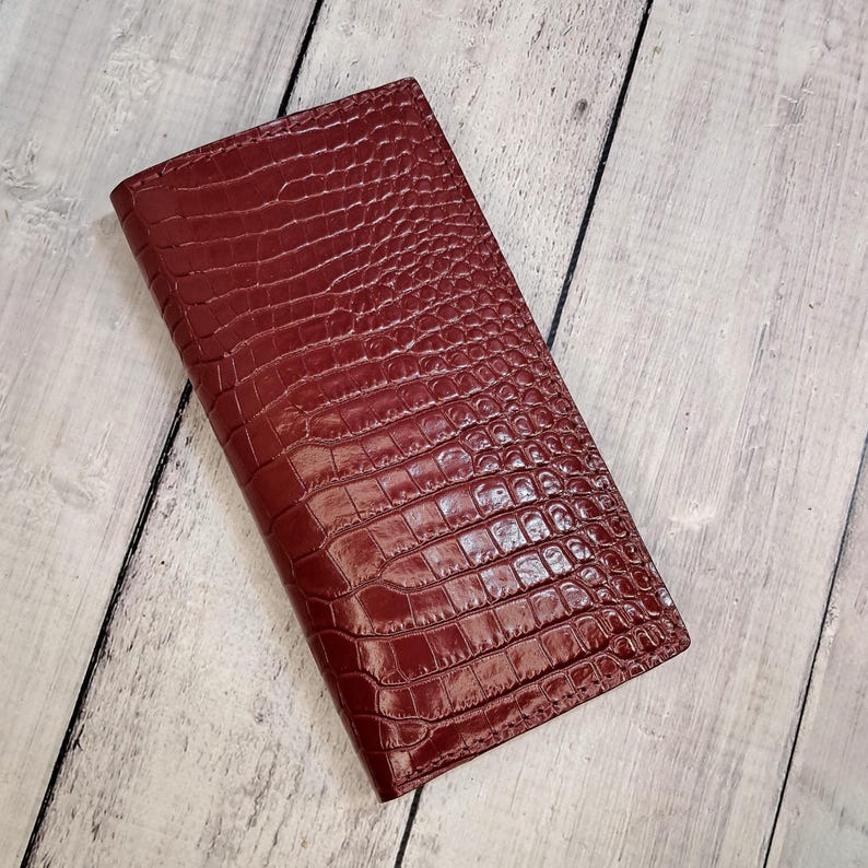 Peut inclure: Un portefeuille rectangulaire d'une couleur bordeaux profond avec un motif textur&eacute; en peau de crocodile. Le portefeuille pr&eacute;sente une finition brillante et une forme fine et allong&eacute;e aux bords droits, avec un motif d'&eacute;cailles gaufr&eacute; uniforme.