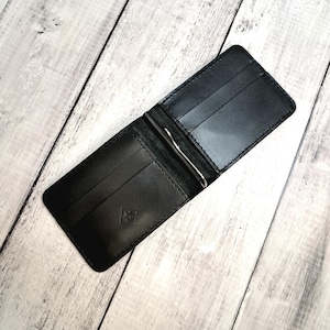 May include: Open black leather wallet with a silver money clip. The wallet has multiple card slots and a small embossed emblem on the front. The wallet is open, displaying the interior compartments.