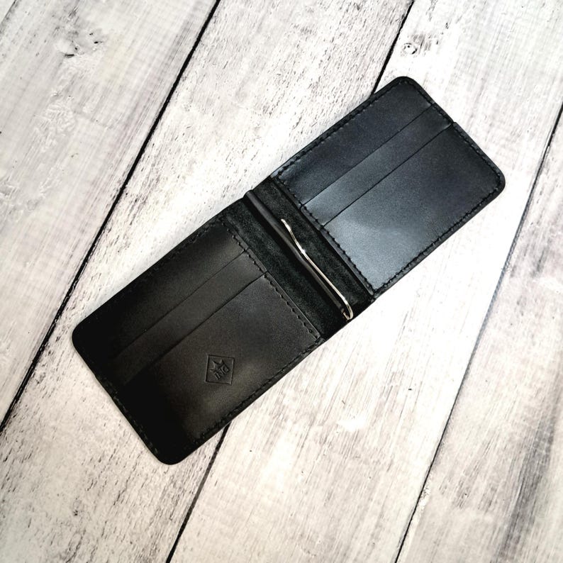 May include: Open black leather wallet with a silver money clip. The wallet has multiple card slots and a small embossed emblem on the front. The wallet is open, displaying the interior compartments.