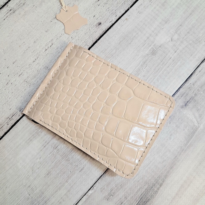 May include: A light beige leather money clip with a crocodile skin texture. The clip is rectangular with a metal clasp and stitched edges. A small leather tag hangs above the clip.
