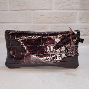 May include: A rectangular, glossy, burgundy-colored clutch with a crocodile skin pattern. The clutch has a zipper closure and a small leather tag attached with a red string. The bag is made of a shiny material.
